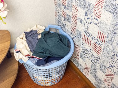 Çeşitli kıyafetlerle dolu mavi bir çamaşır sepeti bir odanın köşesinde duruyor. Sepet, mavi ve kırmızı aksanlı desenli bir duvar kağıdının yanına yerleştiriliyor. Odanın ahşap zemini ve küçük bir ahşap masası var..