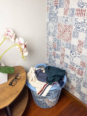 Çeşitli kıyafetlerle dolu bir çamaşır sepeti küçük yuvarlak bir masanın yanına ahşap bir zemine yerleştirilir. Masada gözlük ve saksıda orkide var. Odanın arka planında desenli bir duvar kağıdı ve ahşap bir zemin var. Açık mavi çamaşır sepeti.