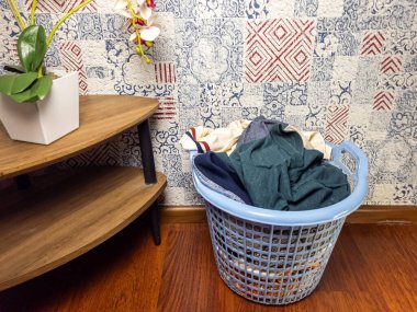 Elbiselerle dolu mavi bir çamaşır sepeti bir odada ahşap bir zeminde duruyor. Arka planda kırmızı, mavi ve beyaz yama tasarımlı dekoratif duvar kağıtları yer almaktadır. Solunda beyaz tencerede bitki olan ahşap bir yan masa var. Genel olarak...