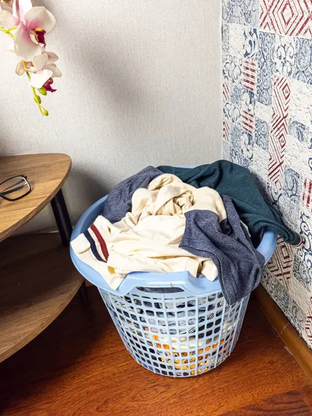 Açık mavi bir çamaşır sepeti çeşitli giysilerle dolup taşıyor. Sepet, beyaz ve desenli duvarları olan bir odanın köşesinde ahşap bir zeminde duruyor. Küçük tahta bir sehpa var, içinde bardaklar ve orkide var..