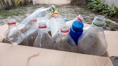 Karton bir kutu geri dönüşüme hazır boş plastik şişelerle dolup taşıyor. Çeşitli boyutlarda ve renkte şişeler görülebilir, bazılarının kapakları hala üzerlerinde. Görüntü geri dönüşümün ve çevresel sorumluluğun önemini vurguluyor. Çanta.