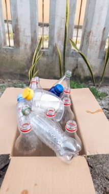 Kullanılmış plastik şişelerle dolu, geri dönüşüme hazır bir karton kutu. Şişeler değişik şekil ve boyutlarda, farklı renkli kapaklarla geliyor. Kutu dışarıda yıpranmış bir duvarın ve bazı bitkilerin karşısında duruyor. Resim bir kıskançlık iletisi iletiyor