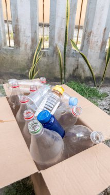 Boş plastik şişelerle dolu, geri dönüşüme hazır bir karton kutu. Çeşitli boylarda ve renklerde şişeler, bazıları kapaklı, kutunun içinde paketleniyor. Sahne açık havada, muhtemelen bir avluda, arka planda çit ve yeşillik var.
