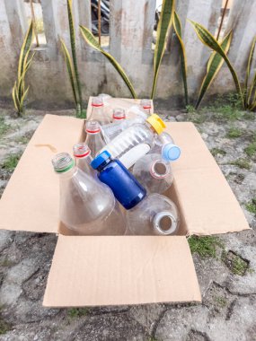 Geri dönüşüme hazır, çeşitli boyutlarda ve şekillerde temiz plastik şişelerle dolu bir karton kutu. Şişeler rastgele kutunun içine konuyor. Bu da çevresel sorumluluğa olan bağlılığını gösteriyor. Kutu dışarıda betonun üzerine konur.