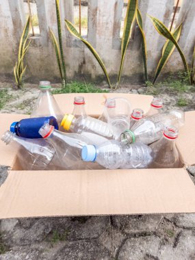 İçinde çeşitli kullanılmış plastik şişeler ve geri dönüşüme hazır plastik bardaklar olan bir karton kutu. Kutu dışarıda, arka planda beton bir duvar ve bitkiler var. Bu görüntü çevresel farkındalığı ve atıkların önemini temsil ediyor. 
