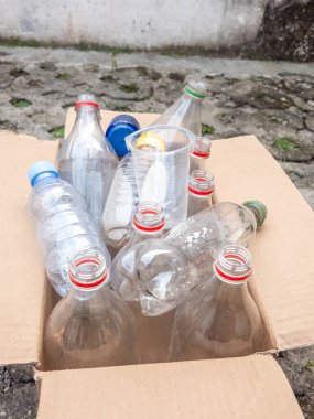 Yakın çekim, çeşitli temiz plastik şişelerle dolu bir karton kutuyu gösteriyor. Şişeler farklı renkte kapaklarla şekil ve boyut olarak farklılık gösterir. Kutu yıpranmış beton bir yüzeyde duruyor. Açık hava yerleşimi ve çevreye odaklanmayı öneriyor.