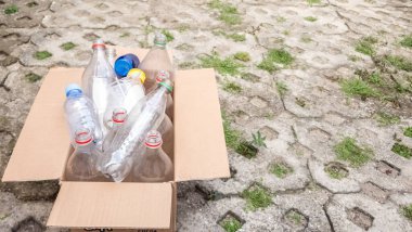 Dışarıda içi plastik şişelerle dolu geri dönüşüme hazır bir karton kutu duruyor. Şişelerin boyutları ve renkleri farklı, mavi, kırmızı ve yeşil şişe kapakları var. Kutu desenli beton bir yüzeye yerleştiriliyor ve çimenler bahislerde büyüyor.