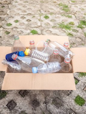 Bir mukavva kutu çeşitli kullanılmış plastik şişelerle ve geri dönüşüme hazır plastik bardakla doludur. Kutu asfalt bir yüzeye yerleştiriliyor ve çatlaklar çimlerle kaplanıyor. Görüntü, çevre koruma ve atık azaltma kavramını vurguluyor