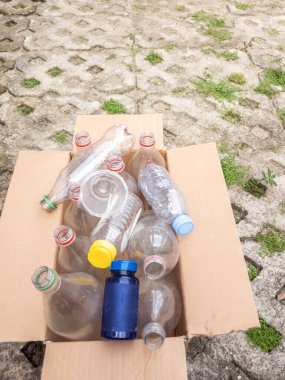 İçinde çeşitli kullanılmış plastik şişeler ve geri dönüşüme hazır mavi bir hap şişesi olan bir karton kutu. Kutu çimenlerle kaplı beton bir yüzeyde duruyor. Çevresel sorumluluk ve atık yönetimine olan bağlılığını gösteriyor. Resim vurguları 