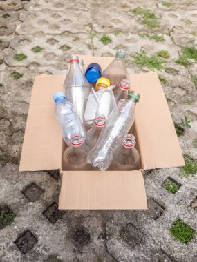 Çeşitli plastik şişelerle dolu bir mukavva kutu beton bir yüzeyde, geri dönüşüme hazır bekliyor. Şişeler farklı boyutlarda ve renklerde, başlıkları hala takılı. Sahne çevresel sorumluluğu ve ithalatı vurguluyor