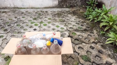 Çeşitli plastik şişelerle dolu, geri dönüşüme hazır bir karton kutu. Kutu, çimen ve bazı bitkilerle desenli, beton bir yüzeyde duruyor. Sahne çevresel farkındalık ve israfın önemini hatırlatıyor.