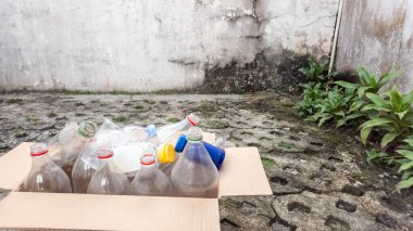 Kullanılmış çeşitli plastik şişelerle dolu bir mukavva kutu dışarıda, geri dönüşüm için hazırlandı. Sahne doğal ışık altında çekiliyor, şişe kapaklarının farklı renklerini ve şeffaf plastiği vurguluyor. Kompozisyon o odaklanır