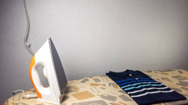 Ütü masasının üzerinde duran ütü desenli mavi çizgili polo tişörtün yakın çekim görüntüsü. Demir turuncu ve beyazdır, solda konumlandırılmıştır. Polo gömleği düzgünce katlanmış. Arka plan düz gri bir duvar. Görüntü günlük d yi yakalar