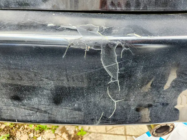 Siyah bir araba tamponuna yakın plan, ciddi bir boya hasarı var. Kabuğu soyulmuş ve çatlak boyalı. Görüntü, aracın dış yüzeyinin yıpranma, yıpranma ve olası çevresel faktörlerin etkilerini gösteren bozulma durumunu vurguluyor. T