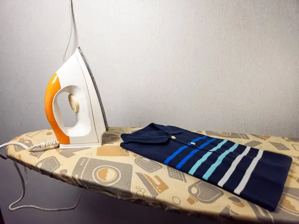 Mavi polo bir gömlek katlanmış ve ütü masasına yerleştirilmiş, ütülenmeye hazır. Tahtada kullanıma hazır bir elektrikli ütü var. Olay yeri kapalı, muhtemelen bir ev ortamı. Işıklandırma yumuşak, evcimen bir hava yaratıyor.