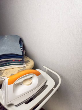 Yakın plan çekimde, yanında katlanmış giysiler bulunan beyaz ve turuncu bir ütü sergileniyor. Kompozisyonu evcimenliği ve ütü yapma eylemini gösteriyor. Görüntü iyi ışıklandırılmış, tarafsız bir arkaplanı var.