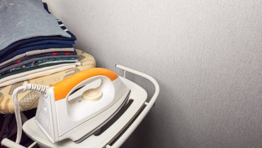 Yakın plan çekimde, bir ütü masasındaki katlanmış kıyafetlerin yanında duran beyaz ve turuncu bir ütü var. Sahne tarafsız gri bir duvara dayanıyor. Görüntü ev ortamını yakalıyor, çamaşırhanenin rutinini vurguluyor.