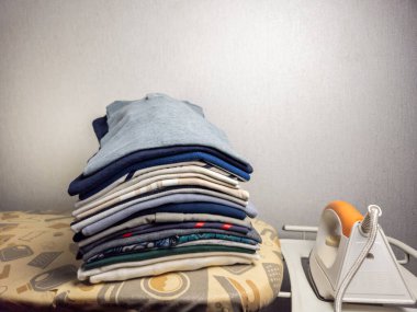 Ütü masasının üzerinde turuncu saplı beyaz ütünün yanında düzgünce katlanmış kıyafetlerin yakın plan çekimi. Arkaplan sade bir duvar, ve görüntü evcillik ve düzen hissi veriyor. Renkler sessiz ve nötr, crea