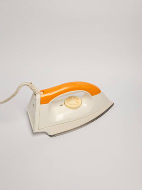 Bu görüntü, turuncu saplı ve ısı kontrol kadranlı beyaz bir vücudun yer aldığı klasik elektrikli ütünün yakın çekimini gösteriyor. Ütü beyaz bir zemin üzerine yerleştirilir ve tasarımı ve detayları vurgulanır. Kompozisyon temiz ve basit.