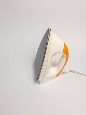 Gri tabanlı beyaz ve turuncu elektrikli demiri yakından çek. Demir düz beyaz bir yüzey üzerinde duruyor, kordon görünür durumda. Görüntü iyi aydınlatılmış ve odak noktası keskin. Kompozisyon basit ve temiz. Reklam için mükemmel.