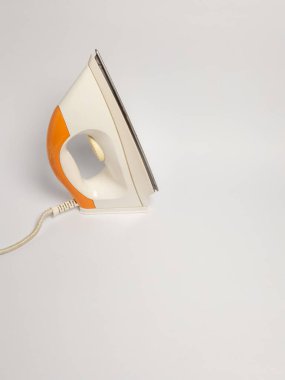 Beyaz bir yüzey üzerinde duran elektrikli ütünün yakın çekim çekimi. Demir, sapı ve kablosu olan turuncu ve beyaz bir şemaya sahiptir. Görüntü temiz, minimalist bir estetik ile çekiliyor, metin veya diğer dergiler için yeterli kopya alanı sunuyor