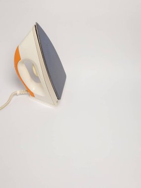 Modern elektrikli ütünün yakın çekim stüdyosu. Ütü, düz beyaz bir arka fona karşı bir açıyla konumlandırılmış. Tasarım, turuncu aksanlı ve gri tabanlı beyaz bir gövdeye sahip. Göbek bağı görünür ve görüntü uygulamayı vurgular.