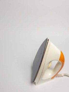 Düz beyaz bir yüzey üzerinde duran bir demiri içeren açılı yakın çekim. Ütü öncelikle beyaz ve turuncudur ve bir güç kablosu görülebilir. Görüntü iyi aydınlatılmış ve temiz. Minimalist bir arkaplana karşı objeye odaklanmış durumda.