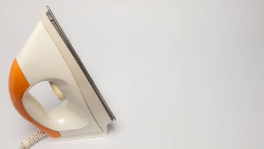 Beyaz arka planda izole edilmiş eski, beyaz ve turuncu bir ütünün yakın plan çekimi. Demir, sapını, taban levhasını ve güç kablosunu gösteren bir açıyla konumlandırılmış. Görüntü ütünün detaylarını vurgular.