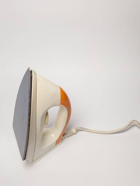 Yakın plan çekimde beyaz ve turuncu renkli retro-stil elektrikli ütü sergileniyor. Demir, düz beyaz bir yüzeye yerleştirilmiştir. Kompozisyon, cihazın tasarımı, şekli ve dokusunu vurgular, sapına ve düz irine vurgu yapar.