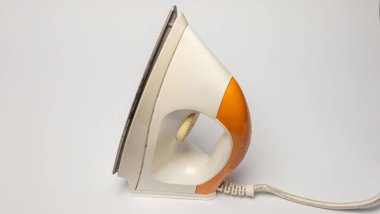 Bu görüntü, beyaz bir yüzeyde duran demiri yakından gösteriyor. Ütü öncelikle beyaz ve sapında turuncu aksanlar var. Güç kablosu görülebilir, demirden uzanıyor. Kompozisyon basittir, nesneyi düzlüğe karşı vurgular. 