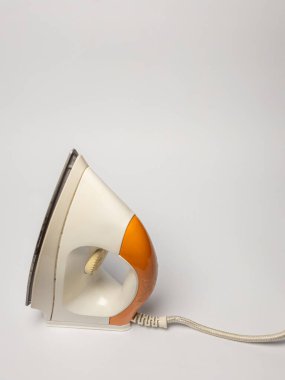 Beyaz arka planda izole edilmiş beyaz ve turuncu elektrikli ütünün yakın plan çekimi. Ütü, tasarımı ve özelliklerini gösteren hafif bir açıyla konumlandırılmıştır. Göbek bağı sağ tarafa doğru uzanıyor ve nesneye bağlam ekliyor. Işıklar... 