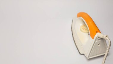 Beyaz arka planda beyaz ve turuncu elektrikli ütünün yakın çekim çekimi. Ütü, tasarımı ve özelliklerini gösteren bir açıyla konumlandırılmıştır. Kapı kolu turuncu, beyaz gövdeyle çelişiyor. Görüntü temiz ve dağınık.,