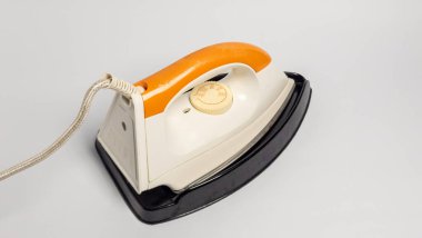 Turuncu saplı ve beyaz gövdeli eski moda elektrikli ütüye yakın çekim, düz beyaz arka plan üzerine kurulmuş. Ütü hafif bir açıyla konumlandırılmış, tasarımı ve özellikleri sergileniyor, sıcaklık arama ve güç kablosu da dahil..