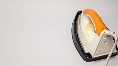 Beyaz bir yüzeyde duran demirin yakın plan çekimi. Ütü, turuncu saplı, beyaz ve siyah bir zeminde duruyor. Güç kablosu görünür durumda. Görüntü iyi aydınlatılmış, temiz, minimalist estetik, ev işlerini göstermek için mükemmel.