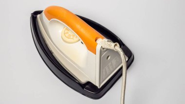 Siyah tabanında duran eski elektrikli ütünün yukarıdan çekimi. Ütüde turuncu bir sapı ve beyaz bir vücudu var. Ütünün güç kablosu görülebiliyor ve kompozisyona işlevsel bir detay ekleniyor. Arka plan... 