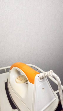 Beyaz bir rafta duran beyaz ve turuncu elektrikli demiri yakından çek. Ütü hafif bir açıyla konumlandırılmış, tasarımı ve detaylarını vurguluyor. Arka plan, demirin ana odak noktası olmasını sağlayan sade açık gri bir duvar. Şey...