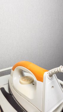 Beyaz ütü masasının üzerinde duran beyaz ve turuncu demiri yakından çek. Ütü çerçevenin içinde, kordon görünür durumda. Arkaplan, tarafsız bir zemin sağlayan düz gri bir duvar. Görüntü ütünün detaylarına odaklanır.