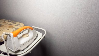 Ütü masasının yakın çekimi ve rafta duran elektrikli ütü. Ütü beyaz, turuncu saplı ve kadranlı. Ütü masası kısmen basılı kapakla görülebilir. Arka plan desenli, gri bir duvar. Işıklı bile.. 