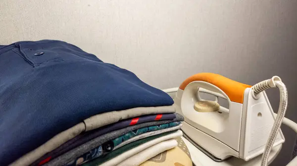 Yakın plan çekimde, beyaz ve turuncu ütüyle birlikte düzgünce istiflenmiş bir yığın kıyafet sergileniyor. Bu da evcilliği gösteriyor. Bu kompozisyon kumaşların berraklığı ve demirin parlak tasarımı üzerine odaklanarak doku ve renkleri vurgular.