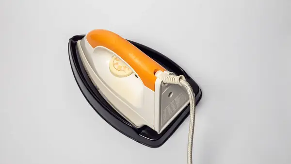 Tepeden çekimde siyah tepsinin üzerinde duran turuncu ve beyaz bir ütü görülüyor. Ütüde sıcaklık göstergesi ve gümüş bir güç kablosu var. Görüntü iyi aydınlatılmış, tarafsız bir ton ve ütünün tasarımına odaklanmış. Bu... 