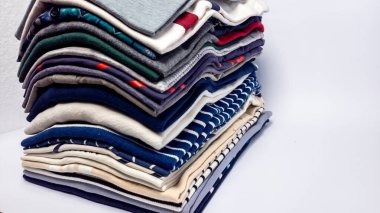 Farklı renk ve kalıplarda düzgünce dizilmiş bir yığın beyaz arka planda duruyor. Giysiler arasında tişörtler, polo tişörtler ve depolanmaya veya sergilenmeye hazır diğer günlük giysiler yer alıyor. Kompozisyon temiz ve minimalist, F ile.