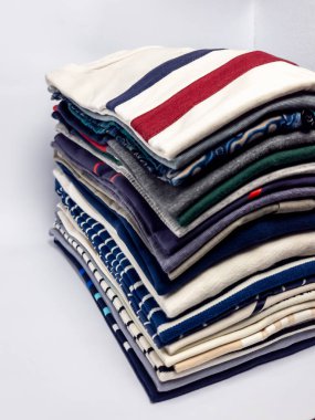 Düzgünce istiflenmiş katlanmış kıyafet yığını, çeşitli renkler ve desenler sergiliyor. Giysiler tişört ve belki birkaç kazak ya da tişört. Görsel olarak çekici bir düzenleme yapıyorlar. Kompozisyon basit, temiz bir beyaz torbayla.