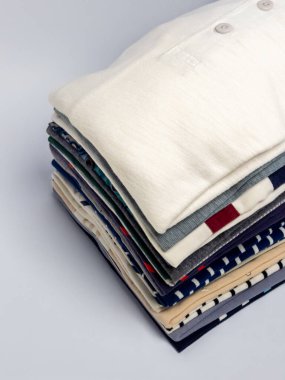 Düzgünce dizilmiş bir yığın katlanmış giysi, çeşitli renk ve desenler sergiliyor. Giysiler beyaz bir yüzeyde, temiz ve minimalist bir estetik yaratıyor. Elbiselerin dokuları ve tasarımları üzerinde duruluyor..