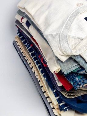 Yakın çekim bir stüdyo sergiler düzgünce istiflenmiş kıyafet öğeleri yığını. Çeşitli renkler, desenler ve desenler içeren çeşitli tişörtler, tişörtler ve muhtemelen diğer giysiler yer alıyor. Giysiler beyaz bir sırt üzerinde düzenlenmiştir.