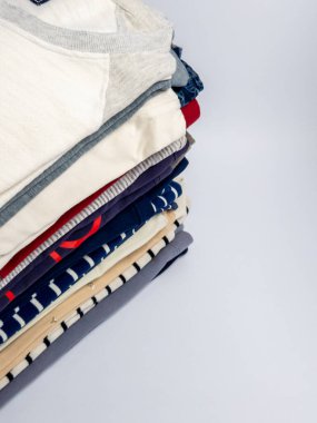 Tişört, kazak ve günlük giysi de dahil olmak üzere düzgünce dizilmiş bir yığın giysinin yakın çekim stüdyosu. Giysiler çeşitli renk ve desenlerde, açık gri bir zemin üzerine yerleştirilmiş, sağda geniş bir kopya alanı var. Th