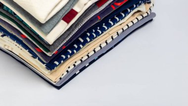 Çeşitli renkler, desenler ve dokulardan oluşan düzgünce istiflenmiş bir kıyafet yığını. Giysiler beyaz bir yüzeyde, temiz ve düzenli bir kompozisyon oluşturuyor. Görüntü, ga 'nın farklı tasarımlarını ve kumaşlarını vurgular.