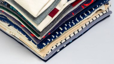 Düzgünce dizilmiş bir yığın katlanmış giysi, çeşitli renk ve desenler sergiliyor. Bileşim açılı, kıyafetlerin farklı katmanlarını ve dokularını vurguluyor. Giysiler temiz, beyaz bir arkaplanda dizilmiş.