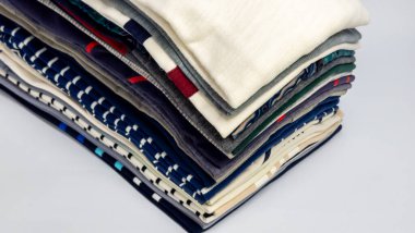 Katlanmış giyim eşyalarının sergilendiği yakın plan bir fotoğraf. Çeşitli desenler ve renklerin yer aldığı giysiler beyaz bir yüzeye yerleştirilmiş ve üst katman krem renkli bir gömlek içermektedir. Işıklandırma dokuları ve ayrıntıları vurgular