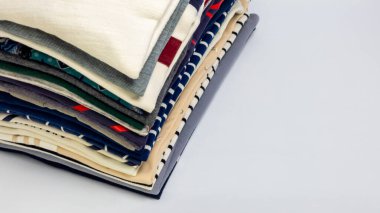 Düz beyaz bir yüzeye düzgünce yerleştirilmiş katlanmış kıyafetlerin yakın plan çekimi. Yığında çeşitli renk ve desenler yer almakta olup, çeşitli kıyafet çeşitliliğini göstermektedir. Kompozisyon, stac 'ın dokusunu ve düzenli yapısını vurgular.