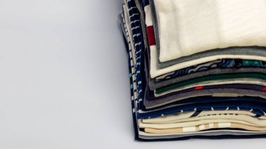 Üst üste düzgünce katlanmış giysiler sergilenen yakın plan bir fotoğraf, ağırlıklı olarak gömlekler, sade beyaz bir arka planda düzenlenmiş. Giysilerin çeşitli renkleri ve desenleri görsel olarak ilginç bir kompozisyon oluşturuyor. Görüntü organizasyonu ve... 